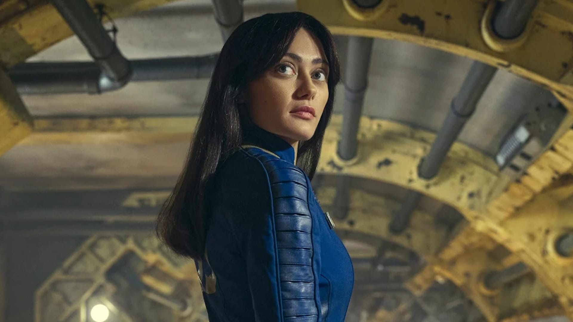 Lucy, wearing a blue vault outfit, looks back above the camera as she is about to leave the vault, full of yellow-painted curved beams and pipes.