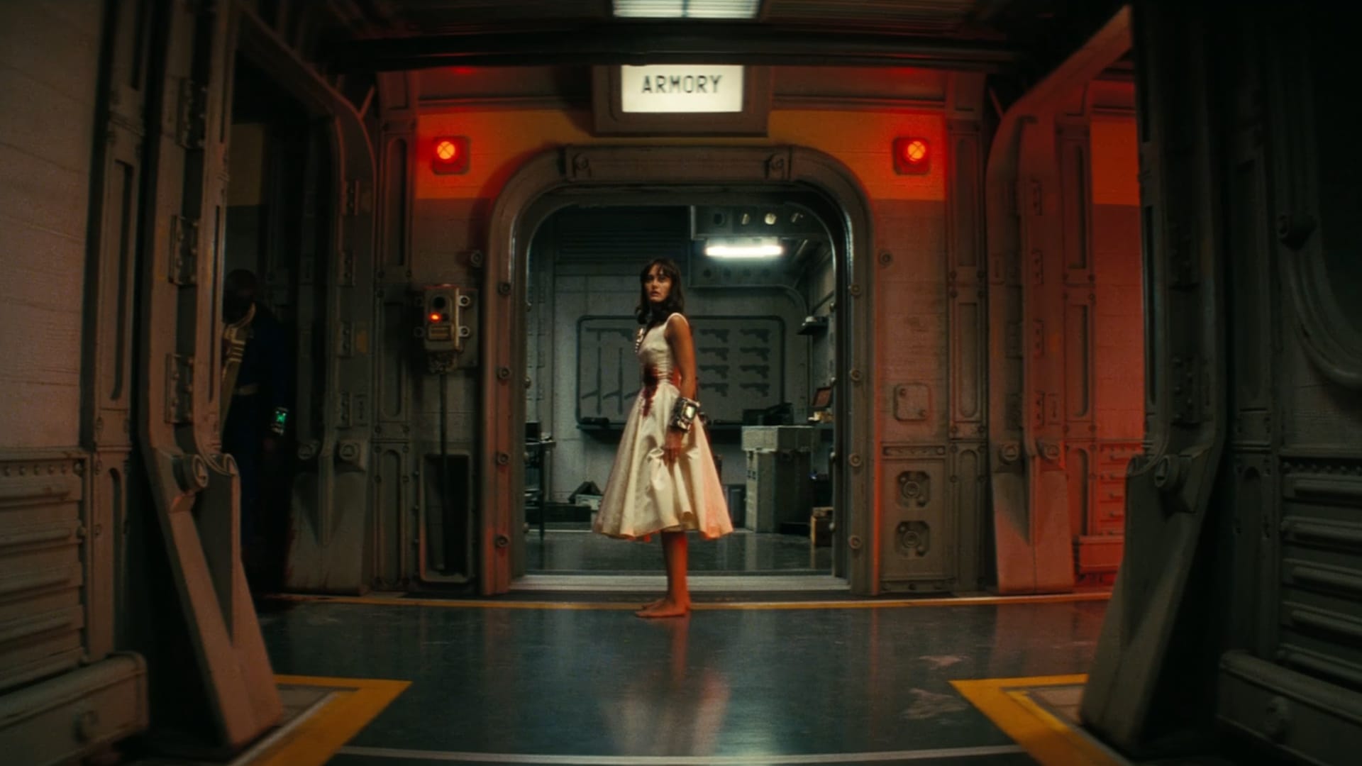 Lucy, barefoot in a white blood-stained dress stares at the camera in front of an open door that says Armory