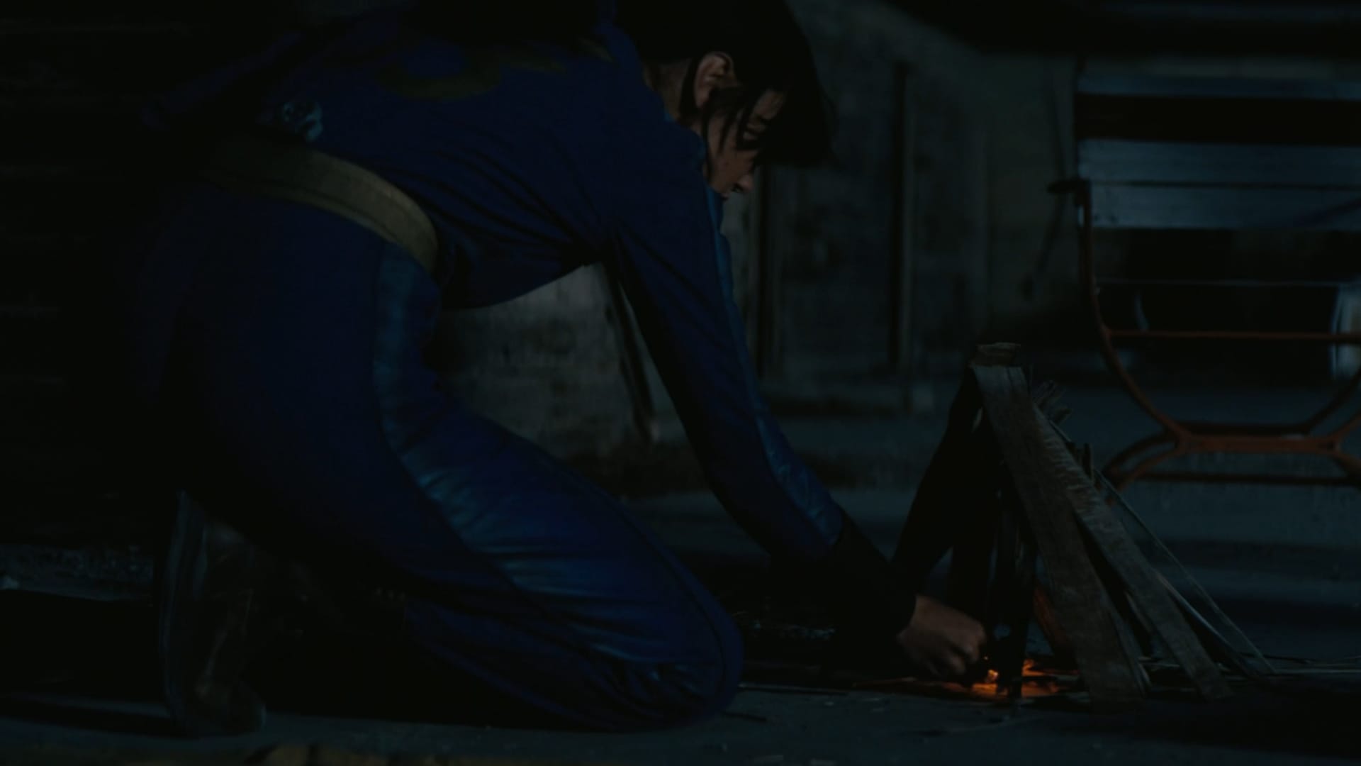 Lucy, in near darkness wearing her full blue fallout shelter regalia, lights a carefully crafted fire with kindling set up in a teepee formation