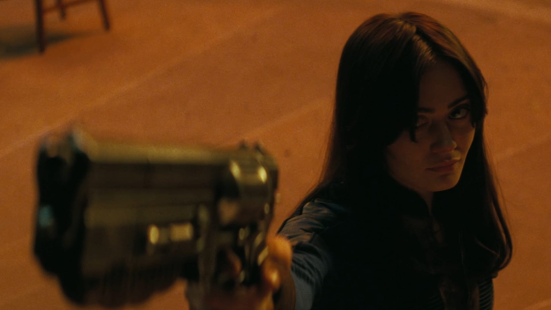 Lucy, wearing her blue vault clothes and with her hair down, points a black metallic gun upward toward the camera with an intense look on her face, against an orange backdrop 